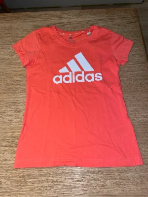 Korallröd Adidas t-shirt med logga - Snygg korallröd t-shirt från Adidas med stor vit logga på bröstet. Klassisk rund halsringning och korta ärmar. Tillverkad i mjuk bomull som känns skön mot huden. Perfekt för en sportig och avslappnad stil.