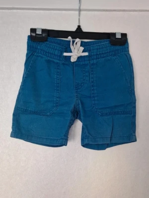 Blå shorts med snörning - Färgen är något blekt efter tvätt, en tråd i tyget på shortsens baksida har gått sönder och det är några ljusa prickar på framsidan av högra benet. Inga shorts till uppklädda tillfällen men passar utmärkt till lek och bus. Stl 98 