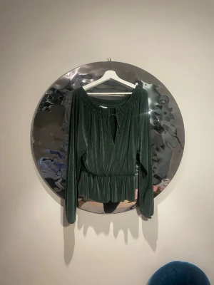 Mörkgrön plisserad blus med knyt - Säljer en mörkgrön plisserad blus med långa ärmar och snygg knytning vid halsen. Blusen har resår i midjan för en skön passform och är gjord i ett mjukt, glansigt material som ger en lyxig känsla. Perfekt att styla med jeans eller kjol.