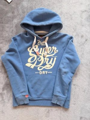 Blå Superdry hoodie med tryck - Säljer en blå hoodie från Superdry med stort vitt tryck på bröstet och klassiska dragsnören i huvan. Hoodien har känguruficka framtill och ribbade muddar vid ärmar och nederkant. Perfekt för en avslappnad streetwear-look.