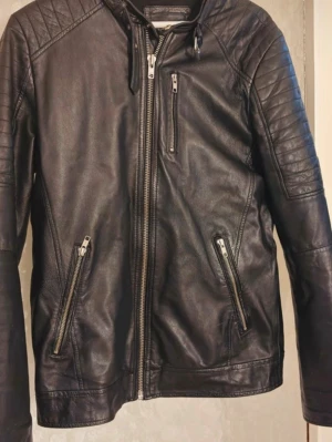 Svart skinnjacka - Snygg svart skinnjacka  och dragkedja framtill. Jackan har quiltade detaljer på axlarna och tre dragkedjeförsedda fickor. Perfekt för dig som vill ha en stilren och cool look. Passar till det mesta och ger en edgy vibe.