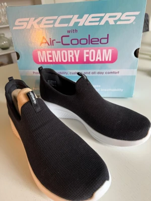 Svarta slip-on sneakers från Skechers oanvända  - Svarta slip-on sneakers från Skechers med vit sula och Air-Cooled Memory Foam för extra komfort. Ovandelen är i meshmaterial som andas och skon har en sportig, stilren look. Perfekta för dig som gillar enkelhet och bekvämlighet. Helt oanvända då det var fel storlek för mig. 