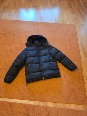 Svart rahlp lauren puffer - Svart pufferjacka från Polo Ralph Lauren med huva och dragkedja framtill. Jackan har en röd broderad logotyp på bröstet, två sidofickor med dragkedja och en bröstficka. Klassisk quiltad design och varm vaddering, perfekt för kalla dagar.