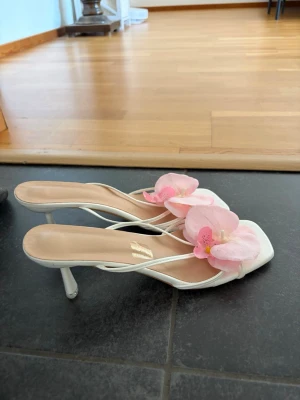 Vita pumps med rosa blomdetalj - Säljer ett par vita pumps med smala remmar och fyrkantig tå. Skorna är dekorerade med rosa blommor framtill. Materialet ser ut att till största del- vara konstläder. Älskar klackarna men passar inte mig längre :/