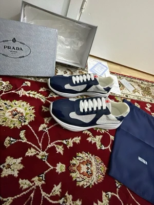 Prada sneakers i blått och vitt - Snygga sneakers från Prada i blått och vitt med klassisk röd logga på plösen. Skorna har en låg silhuett, vita snören och mesh-detaljer för extra andningsförmåga. Perfekta för dig som vill ha en stilren och exklusiv look.