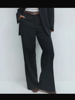 Oanvända kostymbyxor från ginatricot stl S - Produktbild från Gina Tricot för att visa hur byxorna sitter på.  Kritstrecksrandiga kostymbyxor med pressveck framtill. Byxorna är marinblåa med bruna ränder och har medelhög midja. De har dragkedja, bälteshällor, fickor i sidorna och raka ben med normallängd.