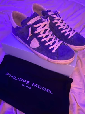 Blå Philippe Model sneakers i mocka - Snygga blå sneakers från Philippe Model i mjuk mocka med vita detaljer och vita skosnören. Klassisk låg modell med vit sula och logotyp på sidan. Perfekta för dig som gillar stilrena och trendiga skor. Säljer dessa skor då jag köpte de och använde ett fåtal gånger under sommaren men sedan har de bara legat på hyllan utan användande. Hör gärna av er om ni har frågor!