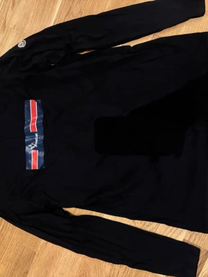 Svart långärmad topp från Moncler - Svart långärmad topp från Moncler med tryckt logga och röd-blå-vit rand på bröstet. Klassisk passform och rund halsringning, perfekt till jeans eller andra byxor. Tidlös design med diskret märke på ärmen.