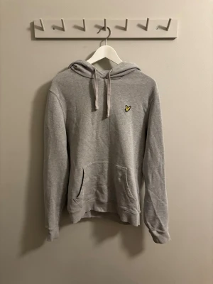 Grå hoodie från Lyle & Scott - Snygg grå hoodie från Lyle & Scott med klassisk gul logga på bröstet. Hoodien har huva med snören och en stor magficka. Perfekt för en avslappnad och stilren look. Köptes för ungefär 2 år sedan men växte ut rätt snabbt så inte använd så mycket. En liten blå prick på magen men syns knappt kan skicka bild om så önskas.