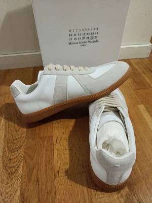 Maison Margiela vita sneakers med mockadetaljer - Maison Margiela sneakers i vitt skinn med beige mockadetaljer och klassisk gummisula. Snygg retrostil med lågt skaft och tonade snören. Perfekta för dig som gillar minimalistisk och stilren design.