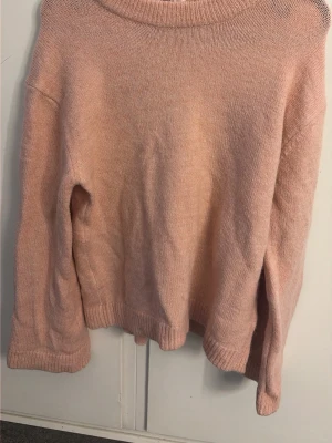 Ljusrosa stickad tröja - Mysig ljusrosa stickad tröja med rund halsringning och långa ärmar. Tröjan har ribbade muddar vid ärmslut och nederkant, vilket ger en avslappnad och skön passform. Perfekt för lager på lager under kyliga dagar.