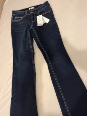 Mörkblå jeans  - Snygga mörkblå bootcut jeans med vita kontrastsömmar och klassiska fem fickor. Jeansen har en ficka bak med lock och knappdetalj samt normalhög midja. Materialet ser ut att vara denim i bomull med lite stretch för skön passform.
