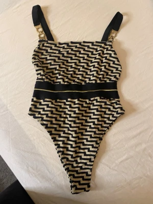 Svart och guld baddräkt med mönster - Snygg baddräkt i svart och guld med zigzag-mönster och bred svart midjedetalj. Axelbanden är svarta med guldfärgade ringar. Perfekt för stranden eller poolen och ger en trendig look.