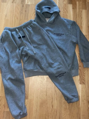 Grå Essentials Fear of God Tracksuit - Snygg grå hoodie och byxor från Essentials Fear of God med svart logga på bröstet. Klassisk passform med huva och känguruficka framtill. Tillverkad i mjuk bomullsblandning som är perfekt för chill dagar. Matchande gråa sweatpants med dragsko och muddar ingår.
