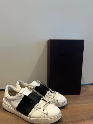 Valentino Opens - Snygga vita sneakers från Valentino Garavani med bred svart läderdetalj över sidan. Klassisk låg modell med rund tå och vita snören. Skorna har en chunky sula och subtila nitar på hälen för extra edge. Medföljer box och Valentino sko snören. Bara att skriva vid några funderingar! 