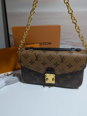 Iouis Vuitton handväska  - Detta är en rektangulär axelremsväska med en klassisk och strukturerad silhuett. Väskan har ett lock som smalnar av till en V-formad flik framtill, vilket ger den ett elegant och symmetriskt uttryck. Material: Väskan är tillverkad i ett mönstrat material med en tydlig textur. Den kombinerar två olika färgställningar: en ljusare brun ovandel på locket och en mörkare brun bas. Mönster: Ytan är prydd med ett genomgående grafiskt mönster bestående av stiliserade blommor och geometriska symboler i en