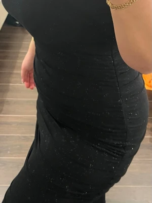 Svart långklänning med glitter - Säljer en svart långklänning med diskret glittrigt mönster. Klänningen har en tight passform och är ärmlös, perfekt för dig som vill ha en elegant och stilren look. Materialet känns mjukt och följsamt.