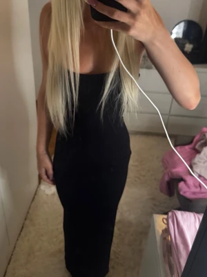 Svart långklänning med smala axelband - En stilren svart långklänning med smala axelband och figurnära passform. Klänningen har en enkel och elegant design som framhäver siluetten. Perfekt för dig som gillar minimalistisk stil och vill ha en klassisk look.