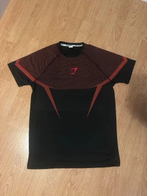 Onyx Gymshark - Snygg svart tränings-t-shirt från Gymshark med röda grafiska detaljer på bröst och rygg. Tillverkad i ett lätt och stretchigt material som andas, perfekt för gymmet eller löpning. Kortärmad modell med rund hals och modern passform.