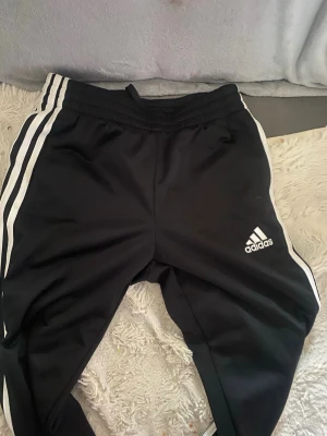 Svarta Adidas mjukisbyxor med ränder - Svarta mjukisbyxor från Adidas med klassiska vita ränder längs sidorna och Adidas-logga på ena benet. Byxorna har resår i midjan och är tillverkade i ett mjukt och bekvämt material. Perfekta för en avslappnad och sportig stil.