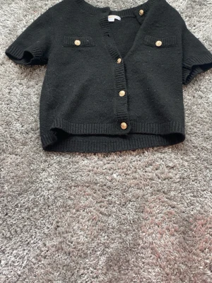 Svart stickad kortärmad kofta Gina Tricot - Kortärmad svart stickad kofta från Gina Tricot med fyra guldfärgade knappar framtill och dekorativa fickor på bröstet. Ribbstickade kanter vid ärmslut och nederkant ger en snygg finish. Perfekt att styla över ett linne eller t-shirt.