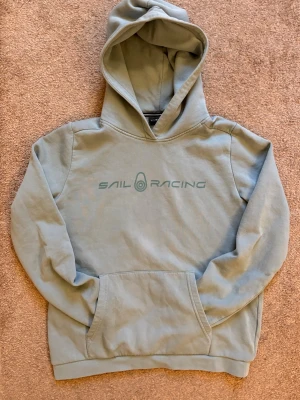 Ljusblå hoodie från Sail Racing - Snygg ljusblå hoodie från Sail Racing med tryckt logga på bröstet. Klassisk känguruficka framtill och stor huva. Tillverkad i mjukt bomullsmaterial som känns skönt mot huden. Perfekt för en avslappnad och sportig stil.