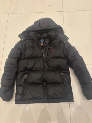 Svart dunjacka från Polo Ralph Lauren - Snygg svart dunjacka från Polo Ralph Lauren med huva och dragkedja framtill. Jackan har två framfickor med dragkedja och den klassiska röda loggan broderad på bröstet. Perfekt för kalla vinterdagar och riktigt skön att bära. Pris kan diskuteras 