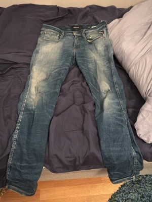 Blå slitna jeans från Replay - Säljer ett par blå jeans från Replay med snyggt slitna detaljer och ljusa partier framtill och baktill. Jeansen har klassisk femficksmodell, dragkedja och knapp i midjan samt en liten reva på höger lår. Perfekta för en avslappnad och trendig look.