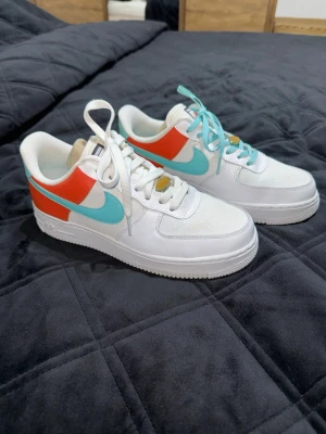 Nike Air Force 1 vita med turkos swoosh - Säljer ett par Nike Air Force 1 sneakers i vitt läder med turkos swoosh och röda detaljer på sidan. Klassisk siluett med perforerad tå och vit sula. Skorna har vita skosnören och en fräsch, sportig look. Använda en gång 