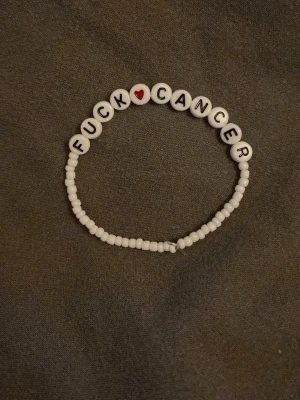 Vitt pärlarmband med text - Ett vitt pärlarmband med bokstavspärlor som bildar texten 'FUCK CANCER' och ett rött hjärta. Armbandet är elastiskt och består av små runda pärlor i plast. Perfekt accessoar för att visa stöd och sprida budskap.