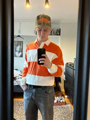 Randig rugbyskjorta i orange och vitt - Säljer en långärmad rugbyskjorta med breda orange och vita ränder, klassisk krage och knappar vid halsen. Skjortan är i bomull och har en loose passform som är riktigt chill. Perfekt för dig som gillar sportig och avslappnad stil.