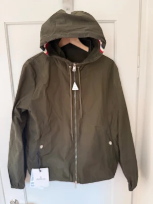 Olivgrön Moncler Grimpeurs Windbreaker - Snygg olivgrön vindjacka från Moncler med huva och dragkedja framtill. Jackan har vita och röda detaljer på huvan, två fickor med tryckknappar och Moncler-logga på ärmen. Perfekt för blåsiga dagar och enkel att matcha med streetwear. Dekaler från ny medföljer. Strl 2/Medium 