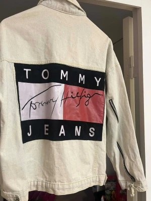 Vit jeansjacka Tommy Hilfiger Vintage 90 tal retro - Snygg vit jeansjacka från Tommy Hilfiger med stort färgglatt logotryck på ryggen och coola dragkedjor längs ärmarna. Klassisk modell med bröstfickor och knappar framtill. Perfekt för dig som vill sticka ut med en unik och trendig jacka. 90 tal Retro, tyvärr är etiketten borta innanför jackan.