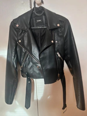 Svart skinnjacka från Pull&Bear - Snygg svart skinnjacka från Pull&Bear med klassisk bikerstil, silvriga dragkedjor, axelklaffar och bälte i midjan. Jackan har flera fickor med dragkedja och en cool, kortare passform som ger en edgy look.