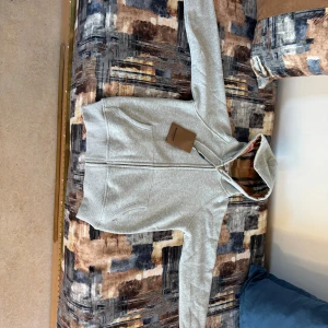 Burberry hoodie  - Snygg grå hoodie från Burberry med dragkedja och klassiskt rutigt foder i huvan. Tröjan har snörning i huvan med metalldetaljer och två fickor framtill. Perfekt för dig som vill ha en stilren och lyxig vardagslook. Obs pytte har gått upp vid fickan (märks ej)