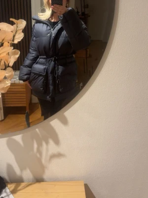 Svart dunjacka från Arket  - Snygg svart pufferjacka med dragsko i midjan för en fin passform. Jackan har två stora fickor framtill och stängs med dragkedja. Helt oanvänd 