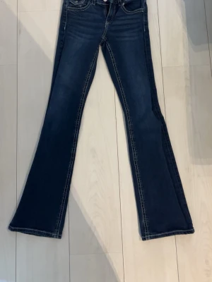 Mörkblå bootcut jeans med kontrastsömmar - Snygga mörkblå bootcut jeans med tydliga kontrastsömmar och klassiska fem fickor. Jeansen har utsvängda ben och låg midja, perfekta för en trendig och avslappnad look. Materialet är denim med stretch för extra komfort.
