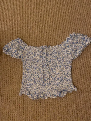 Blommig off shoulder topp blåvit - Supersöt off shoulder topp i blått och vitt med småblommigt mönster och volangkant. Tunn och stretchig, perfekt till sommaren. Korta ärmar och smockad passform ger en trendig och bekväm look.