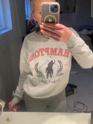 Grå sweatshirt med Hampton Polo Club-tryck - Säljer en grå sweatshirt med stort Hampton Polo Club-tryck i rött och svart på bröstet. Tröjan har rund hals, ribbade muddar och en avslappnad passform. Perfekt för dig som gillar sportiga och klassiska college-vibes.