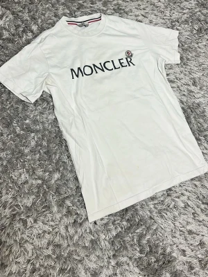 Moncler t-shirt  - Snygg vit t-shirt från Moncler med stor svart logga och emblem på bröstet. Klassisk passform och rund halsringning. Tillverkad i mjuk bomull som känns skön mot huden. Perfekt för dig som gillar stilrena och exklusiva plagg.