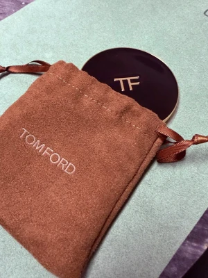 Tom Ford sminkspegel i guld - Lyxig rund sminkspegel från Tom Ford med elegant guldfärgad ram och mörkbrun framsida med TF-logga. Kommer i en mjuk brun sammetsliknande påse med dragsko och broderad logga. Perfekt att ha i väskan för snabba touch-ups.
