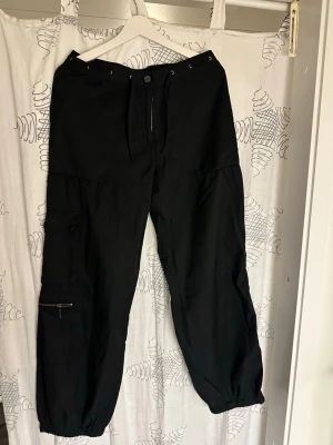 Svarta cargobyxor med dragkedja - Svarta cargobyxor med flera fickor och dragkedja på ena benet. Byxorna har resår vid bensluten och knäppning i midjan. Perfekta för en avslappnad och streetinspirerad stil.