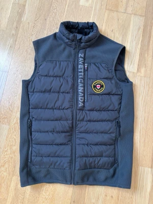 Grå/svart dunväst från Zavetti Canada - Snygg svart dunväst från Zavetti Canada med quiltad design och hög krage. Västen har dragkedja framtill, två sidofickor med dragkedja och ett broderat märke på bröstet. Materialet är en mix av glansig och perforerad textil för en modern look.