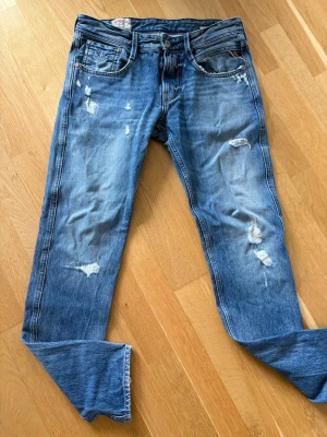 Ljusblå slitna jeans från Replay - Säljer ett par ljusblå jeans från Replay med snygga slitningar och detaljerade sömmar. Jeansen har klassisk femficksmodell, knappgylf och läderpatch bak i midjan. Perfekta för en avslappnad och trendig look.