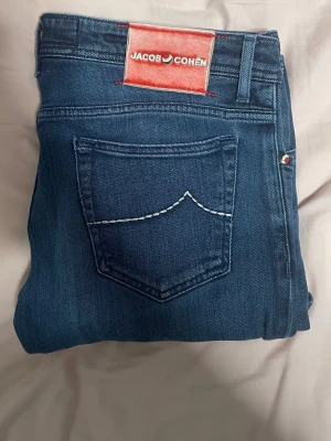 Mörkblå jeans från Jacob Cohen - Snygga mörkblå jeans från Jacob Cohen med klassisk femficksdesign och kontrastsöm på bakfickan. Jeansen har en röd patch med logga bak i midjan och är tillverkade i mjukt denimtyg som ger en skön passform. Perfekta för en trendig och avslappnad stil.