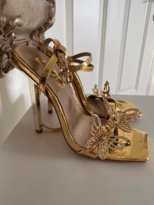 Guldiga klackar - Unika guldiga klackar från EGO med fyrkantig tå, metallic finish och krokodilpräglat mönster. Dekorerade med snygga metallfjärilar och pärldetaljer framtill. Smala remmar och hög klack ger en trendig look som sticker ut.