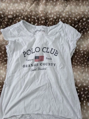 Vit t-shirt från Saint Vincent - Vit t-shirt från Saint Vincent med tryck 'Polo Club Orange County' och amerikansk flagga på bröstet. Klassisk rund hals och korta ärmar. Tillverkad i mjuk bomull, perfekt för en avslappnad stil.