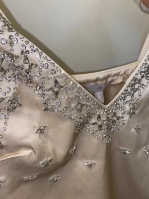 Champagnefärgad klänning med pärlor H&M - Elegant champagnefärgad klänning från H&M med v-ringning och breda axelband. Klänningen är dekorerad med glittrande pärlor och stenar längs urringningen och framtill. Materialet har en satinliknande finish som ger en lyxig känsla. Kan hämtas upp i Stockholm annars kostar frakt extra‼️