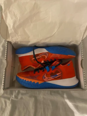 Nike Kyrie Flytrap V sneakers orange/blå - Nike Kyrie Flytrap V sneakers i stark orange och blå med svarta detaljer. Skorna har snörning, vit mellansula och mönstrad blå yttersula. Perfekta för träning och basket, med sportig design och bekväm passform.