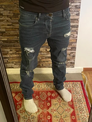 Mörkblå ripped jeans herr - Säljer ett par mörkblå jeans med slitna detaljer och hål på båda benen. Jeansen har en klassisk femficksmodell och normal passform. Perfekta för en avslappnad och trendig streetstil.